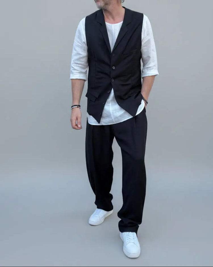 Person wearing a black vest over a white shirt with dark pants and white shoes, in front of light grey backdrop.
