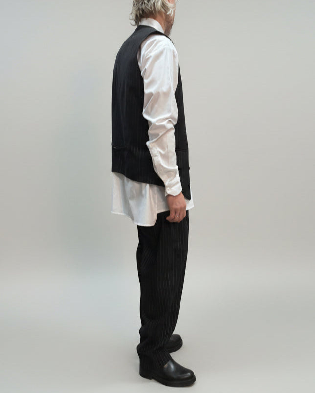 MODEL FACING ON THE SIDE WEARING BLACK PINTRIPE VEST AND LOOSE PANTS IN A RELEXED POSITION