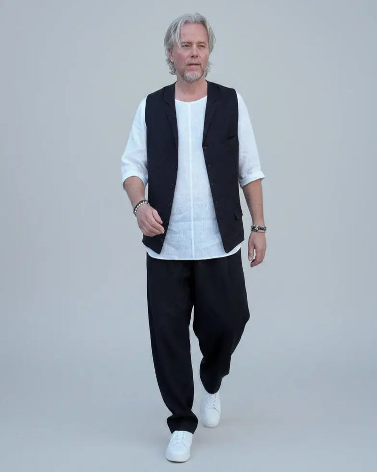 Person wearing a black vest over a white shirt with dark pants and white shoes, in front of light grey backdrop.
