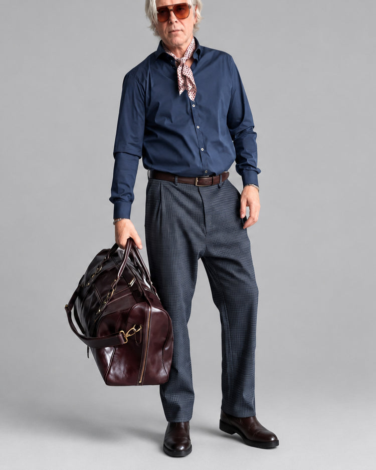 A man wearing a navy blue button-down shirt, patterned neck scarf, and blue?gray micro-check trousers stands in front of a white brick wall, holding a dark burgundy leather duffel bag.
