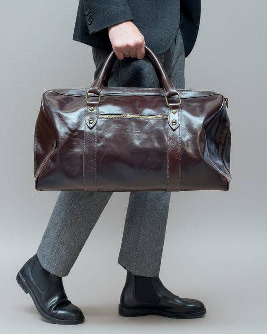 Person holding a brown leather duffel bag with shoulder strap and one puller on front pocket against a plain background