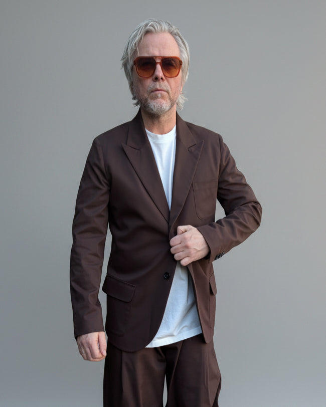 Man in a brown suit and sunglasses standing on a street in front of light grey backdrop