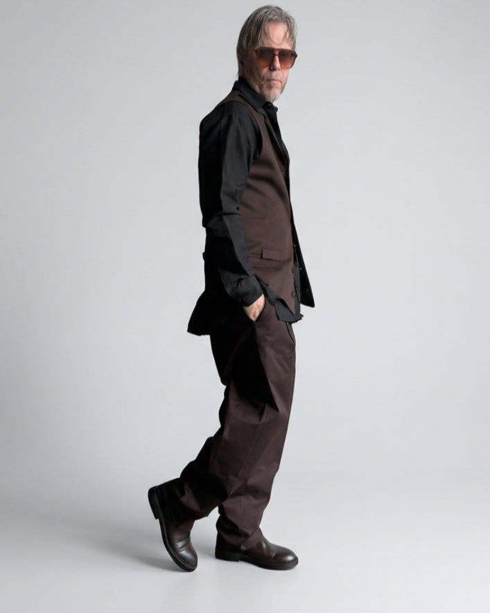 A man wearing a dark brown tailored vest and balloon trousers, black shirt, and sunglasses stands against a white painted brick wall.