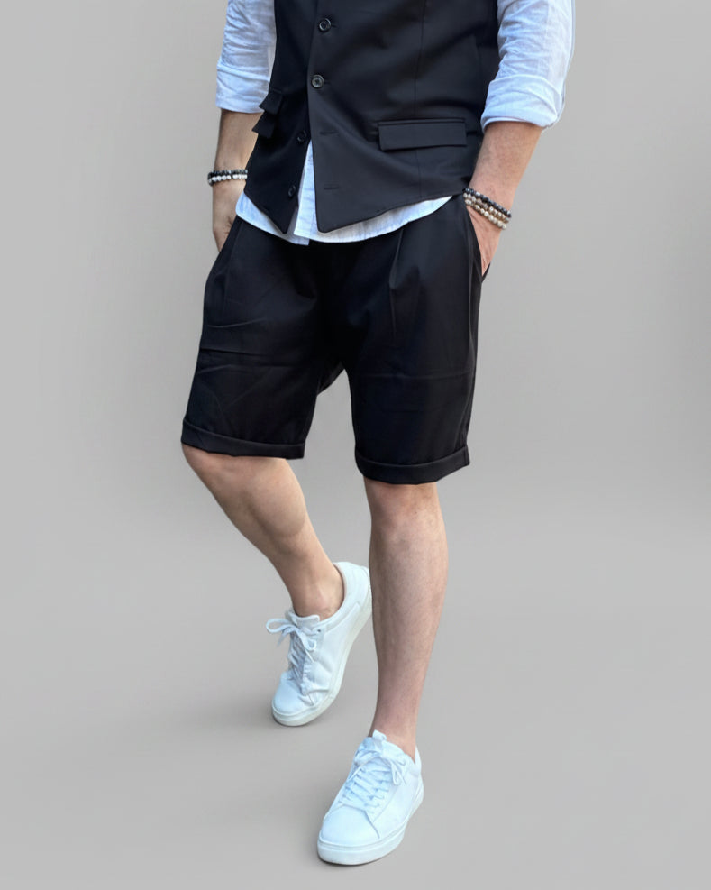 Model in front of light grey backdrop wearing black vest and shorts and white shirt. with tanned legs.