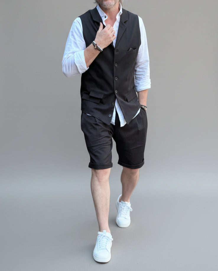 Model in front of light grey backdrop wearing black vest and shorts and white shirt. with tanned legs.