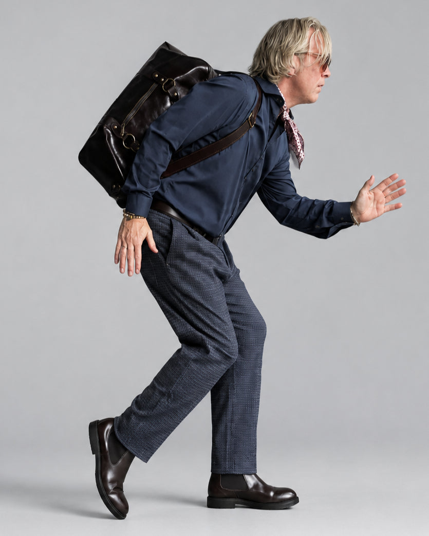 A man in a navy blue shirt, gray/blue micro checked patterned trousers, and brown leather boots is captured mid-stride, leaning forward with his hands outstretched. He is wearing a dark brown leather messenger bag slung over his shoulder and has a patterned neck scarf.