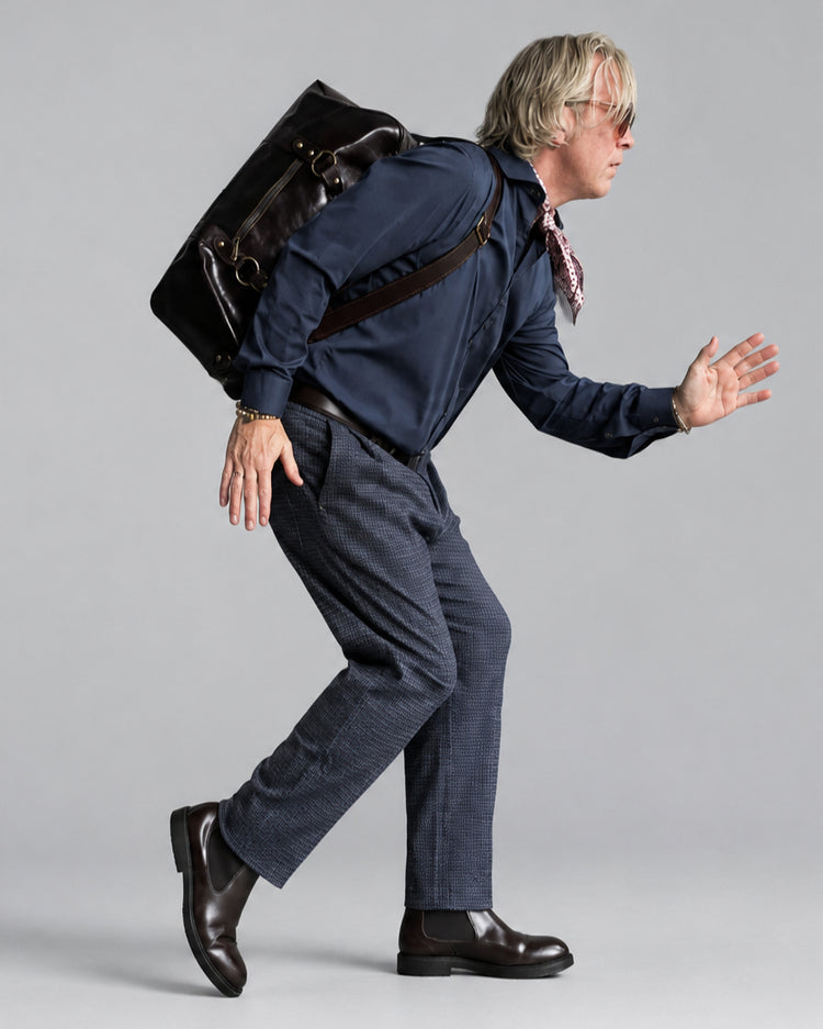 A man in a navy blue shirt, gray/blue micro checked patterned trousers, and brown leather boots is captured mid-stride, leaning forward with his hands outstretched. He is wearing a dark brown leather messenger bag slung over his shoulder and has a patterned neck scarf.