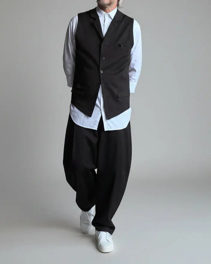 Model in baggy black pants, white shirt and black waistcoat standing in front of light grey backdrop