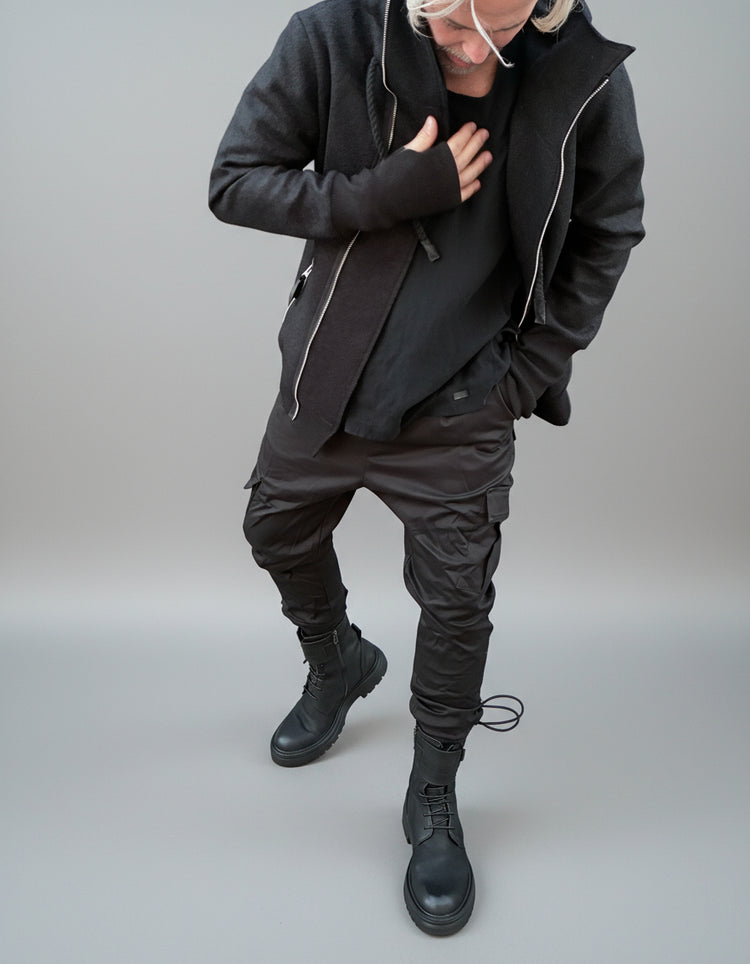 Person wearing a dark outfit and black boots against a gray wall. keep position as it is.