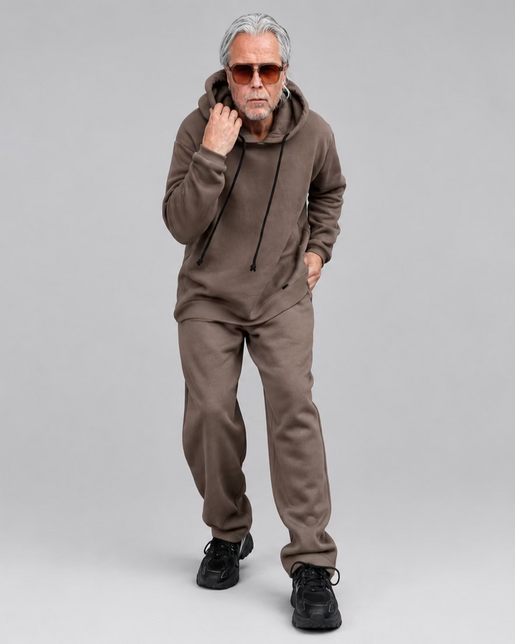 A man wearing a matching brown lounge set consisting of a popover hoodie with no pocket on and sweatpants, standing in front of a black metal door framed by white brick walls.