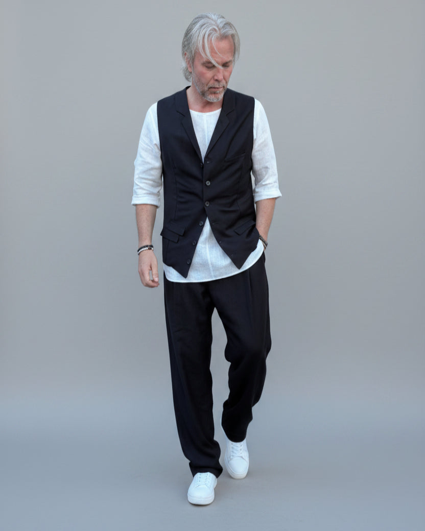 Person wearing a black vest over a white shirt with dark pants and white shoes, in front of light grey backdrop.