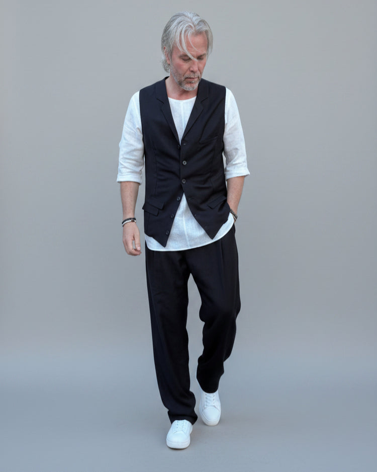 Person wearing a black vest over a white shirt with dark pants and white shoes, in front of light grey backdrop.
