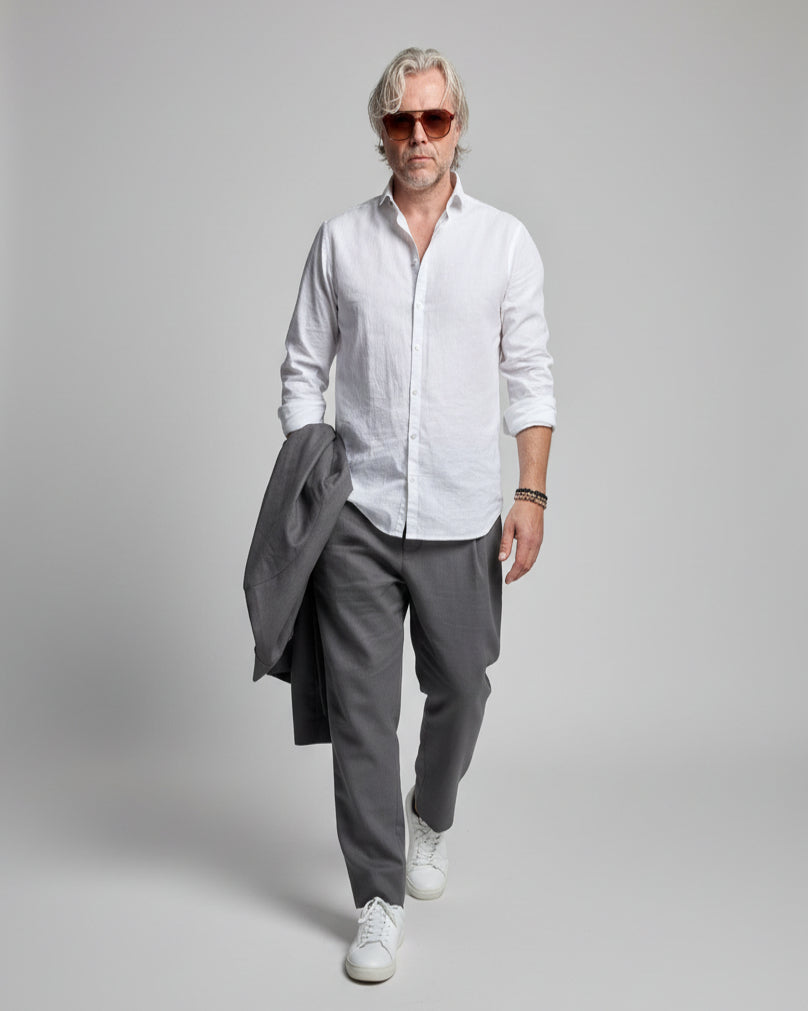 Person wearing a white shirt, gray linen pants, and white sneakers on a light grey background in a dynamic possition.