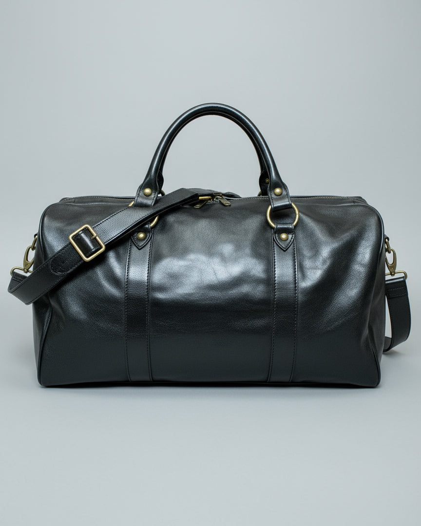 Black leather duffel bag with a shoulder strap and 1 puller on the side pocket on a textured light grey surface