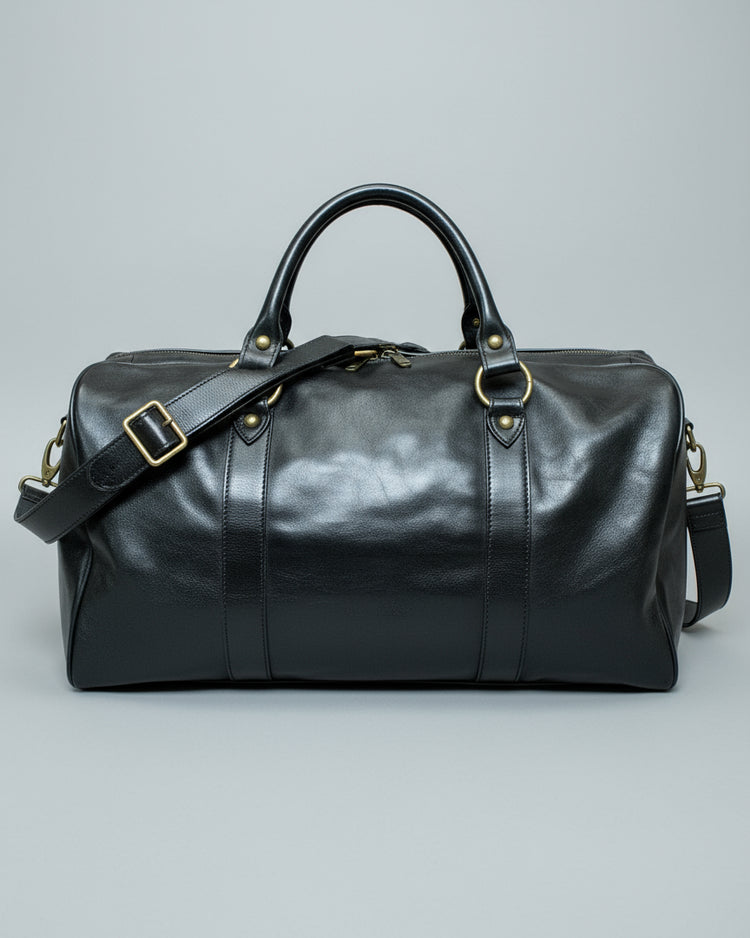Black leather duffel bag with a shoulder strap and 1 puller on the side pocket on a textured light grey surface