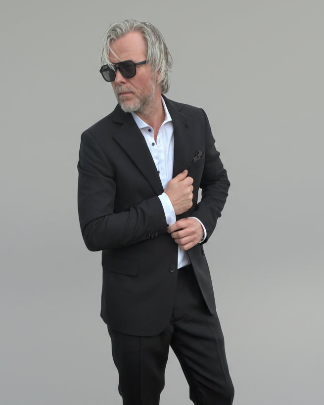 Model standing in front of light grey backdrop wearing black suit and white shirt.