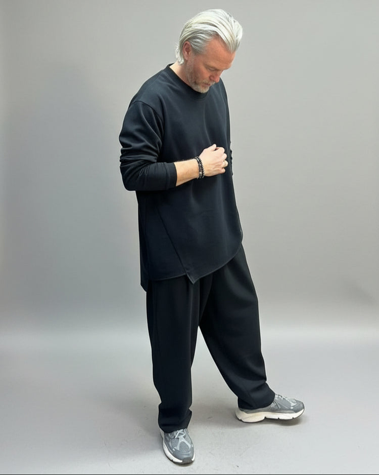 Man wearing a black outfit standing in front of beige backdrop 