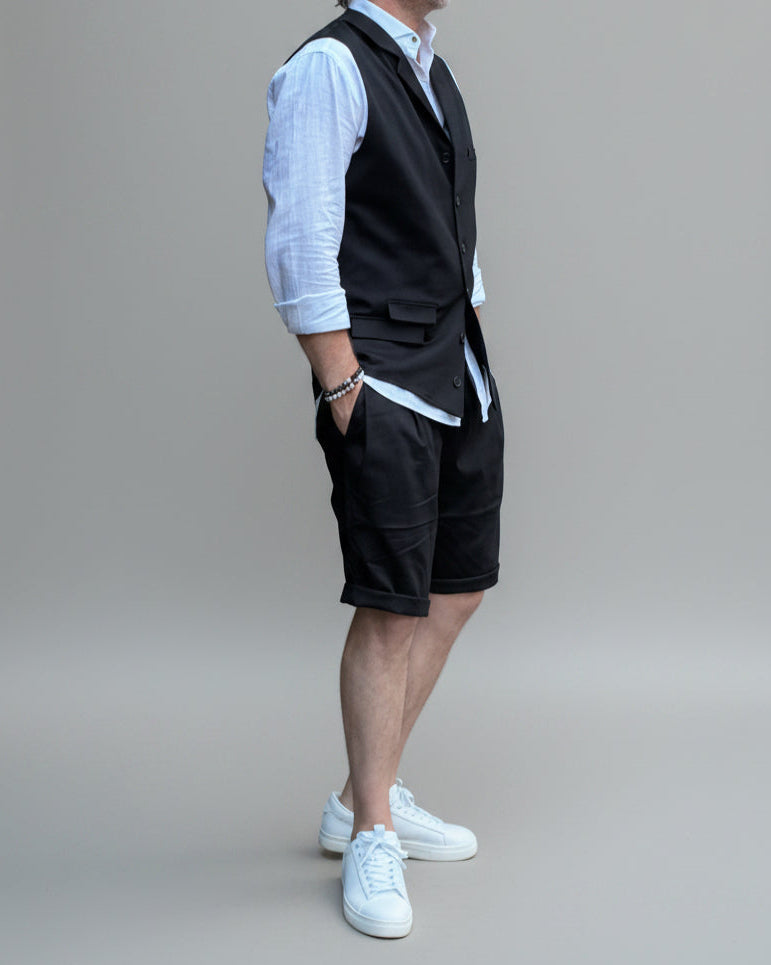 Model in front of light grey backdrop wearing black vest and shorts and white shirt. with tanned legs.