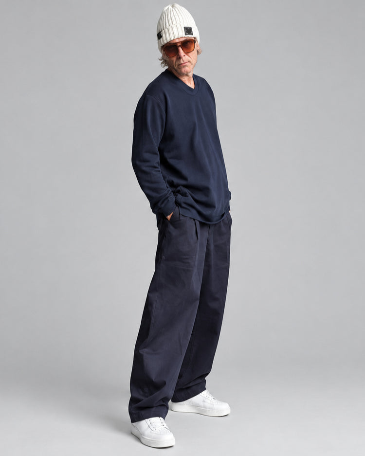 A man wearing a navy blue crewneck sweatshirt, navy pants, white sneakers, and a white knit beanie with orange-tinted sunglasses, standing in front of a white brick wall.