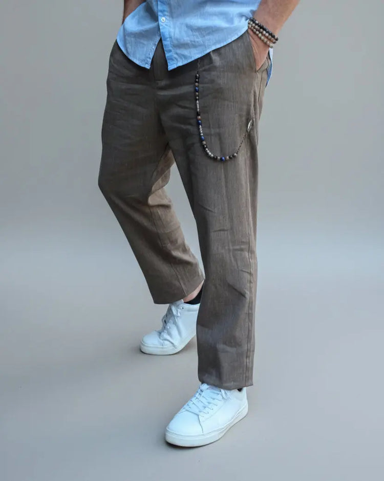 Model standing in front of light grey backdrop wearing brown linen pants and white shirt and sneakers