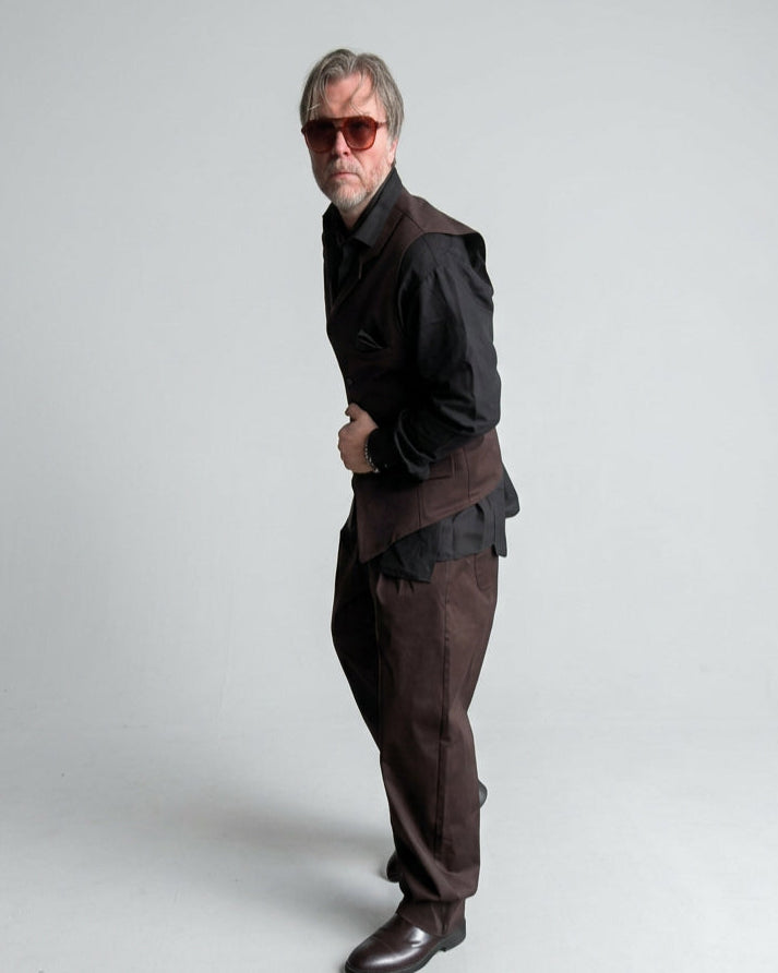 A man wearing a dark brown tailored vest and balloon trousers, black shirt, and sunglasses stands against a white painted brick wall.
