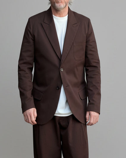 Model wearing brown oversized suit with white shoes and T-shirt in front of light grey backdrop.