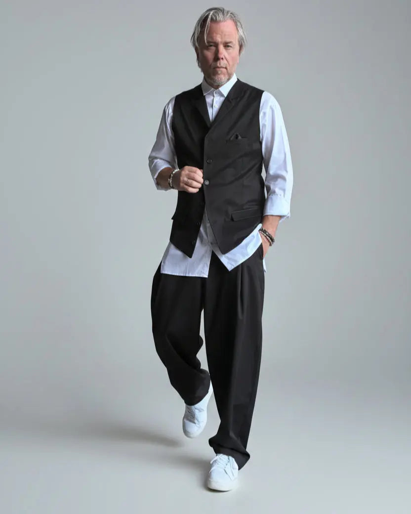 Model in baggy black pants, white shirt and black waistcoat standing in front of light grey backdrop