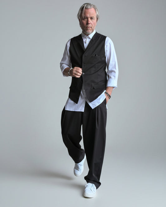 Model in baggy black pants, white shirt and black waistcoat standing in front of light grey backdrop