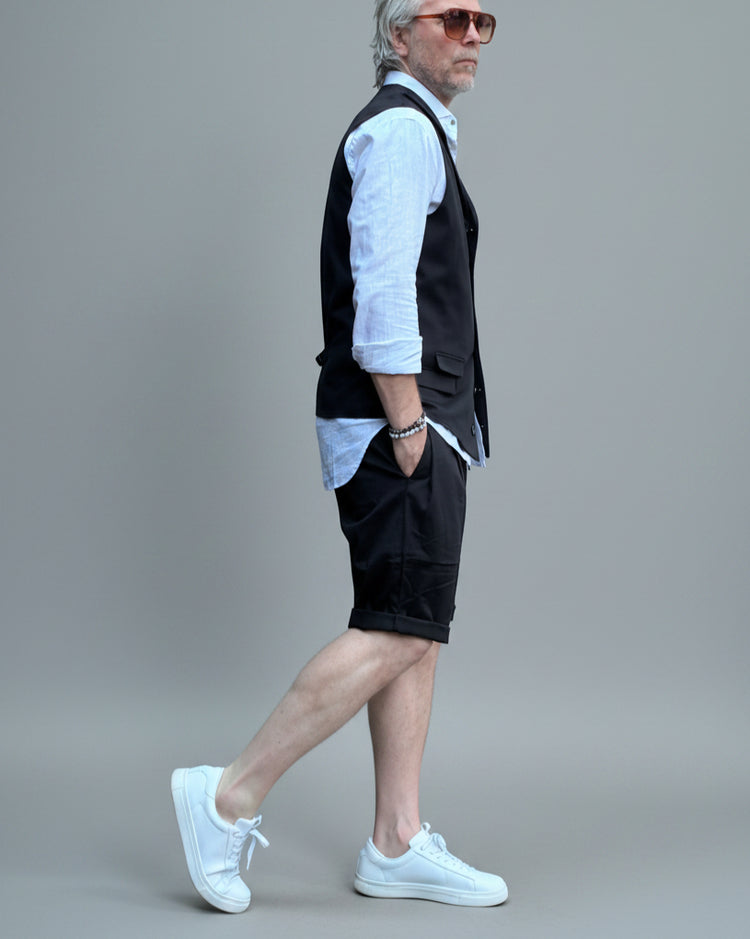 Model in front of light grey backdrop wearing black vest and shorts and white shirt. with tanned legs.