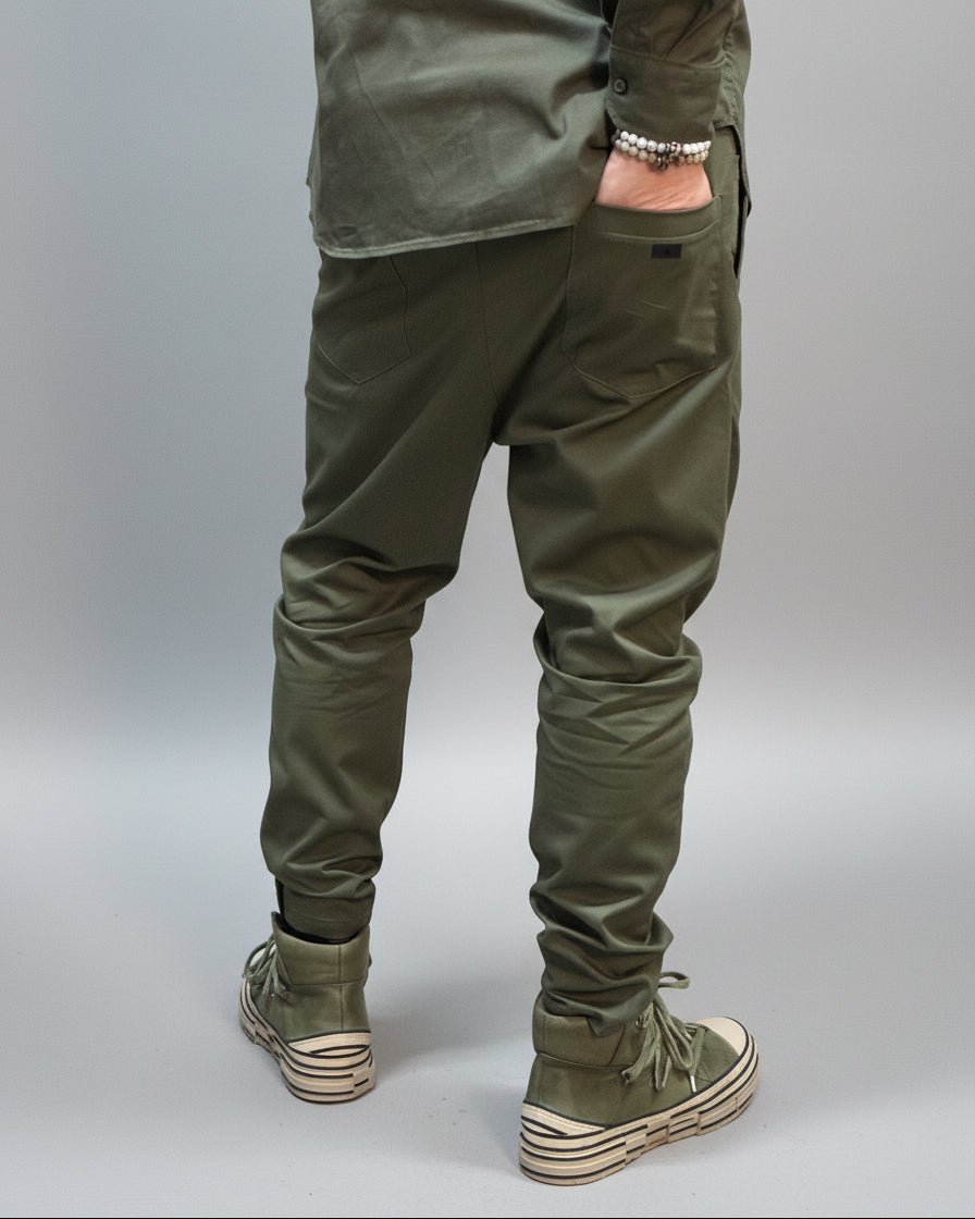 Person wearing green pants and shoes in front of a black background with his right hand in the back pocket.