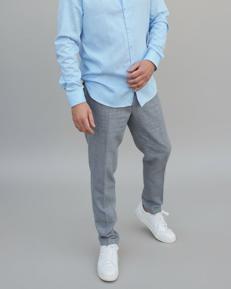 Man in a gray, blue checked pants and light blue shirt standing in front of a light grey backdrop.