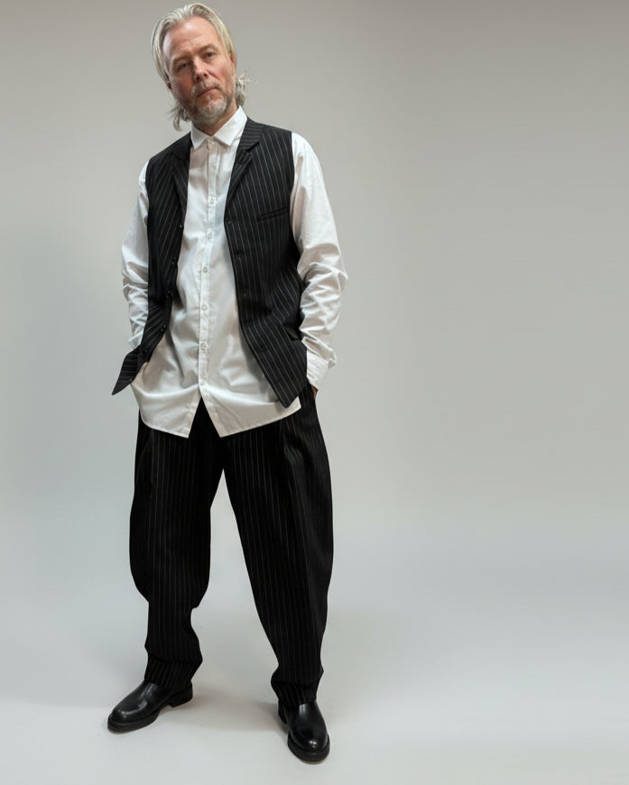 MODEL FACING THE CAMERA WEARING BLACK PINTRIPE VEST AND LOOSE PANTS IN A RELEXED POSITION