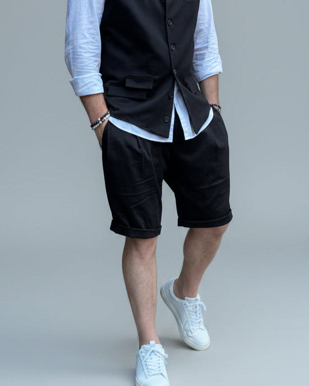 Model in front of light grey backdrop wearing black vest and shorts and white shirt. with tanned legs.