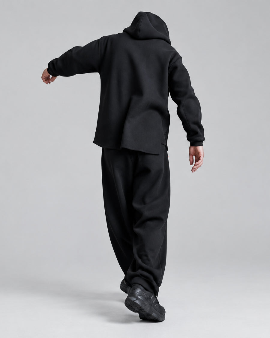 A person wearing a black oversized hoodie and matching black sweatpants, photographed from behind in an outdoor urban setting with a white brick wall and dark doorway behind them.