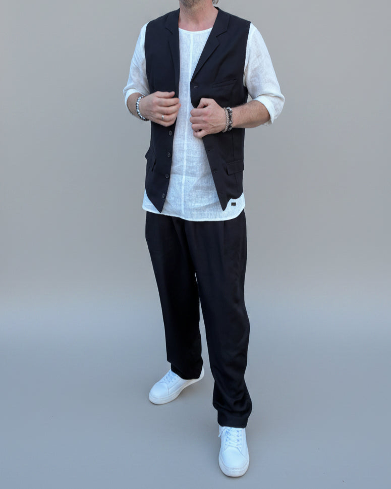Person wearing a black vest over a white shirt with dark pants and white shoes, in front of light grey backdrop.
