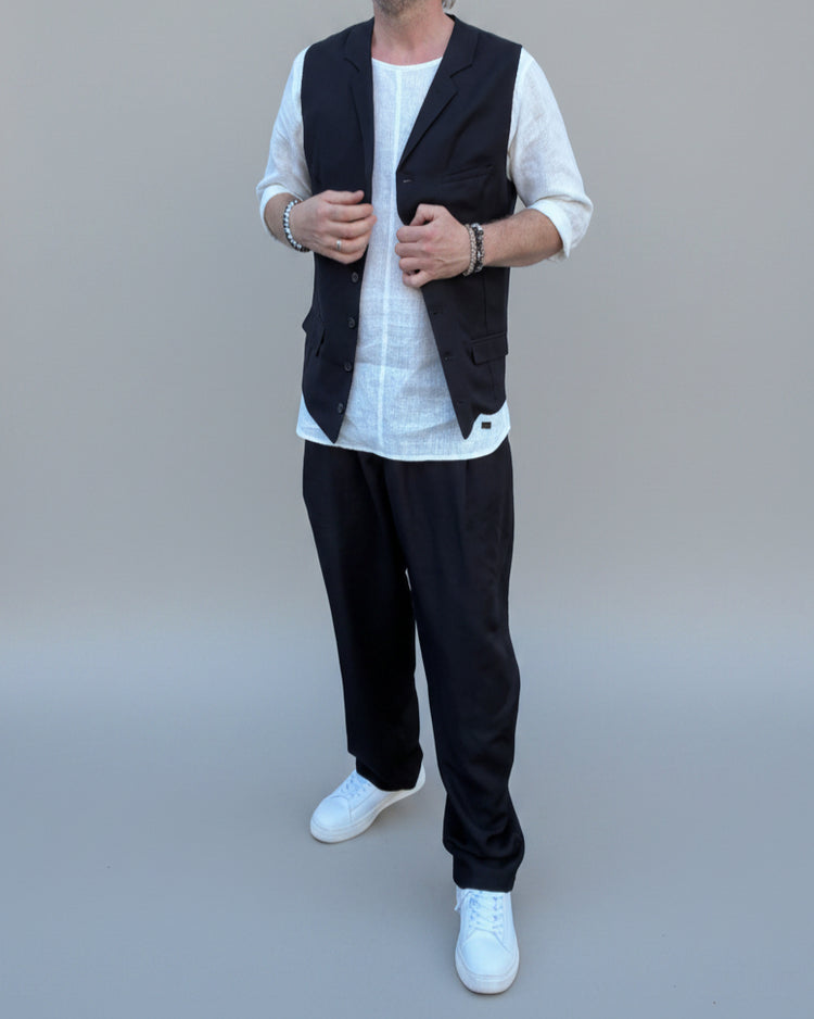 Person wearing a black vest over a white shirt with dark pants and white shoes, in front of light grey backdrop.
