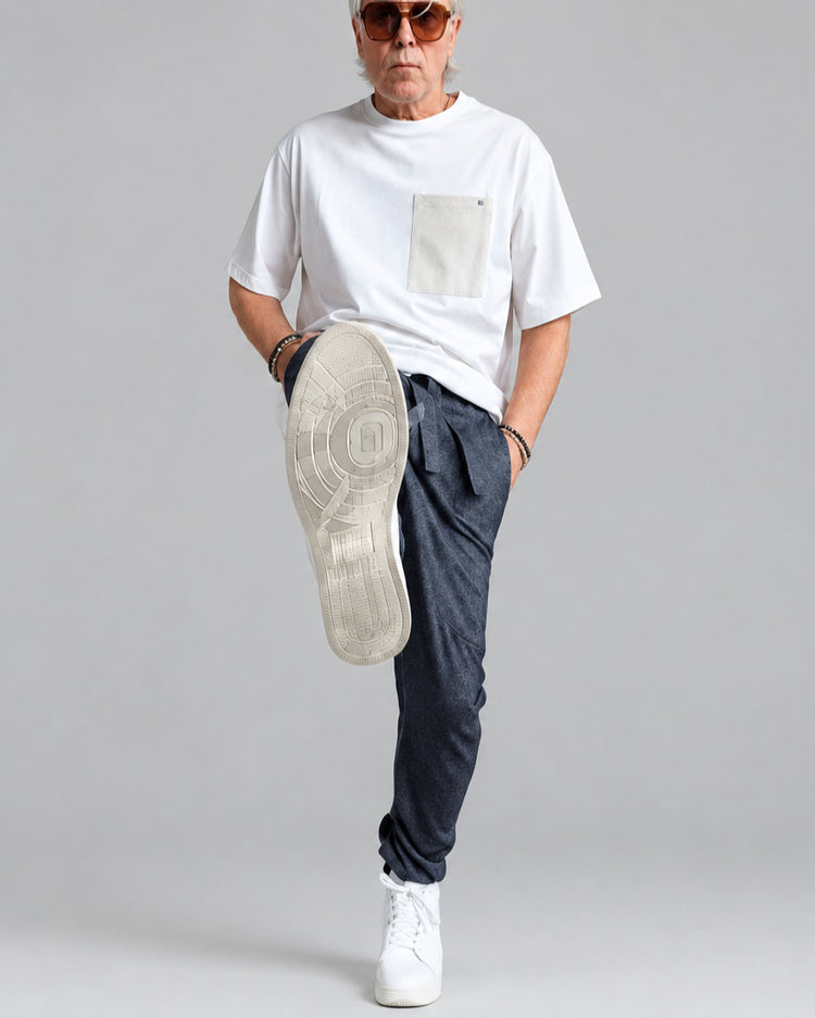 A man wearing a white t-shirt with beige pocket, dark patterned  wool jogger pants, and white sneakers kicks up his left leg to show the sole of a beige sneaker in motion against a white brick wall background.