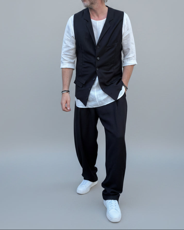 Person wearing a black vest over a white shirt with dark pants and white shoes, in front of light grey backdrop.