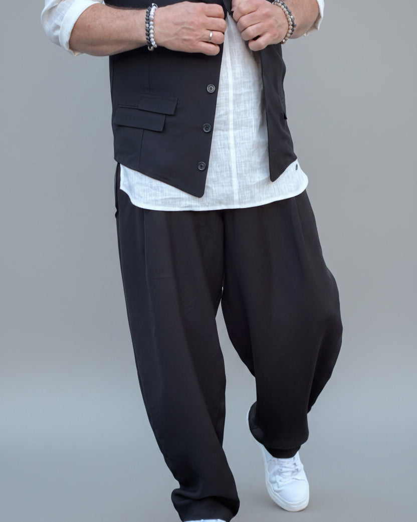 Person wearing a black vest over a white shirt with dark pants and white shoes, in front of light grey backdrop.