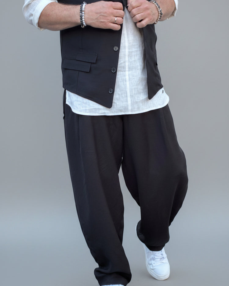 Person wearing a black vest over a white shirt with dark pants and white shoes, in front of light grey backdrop.
