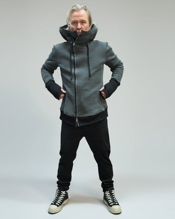 Person wearing a gray hoodie and black pants standing against a light grey curtain holding pullers on zippers.