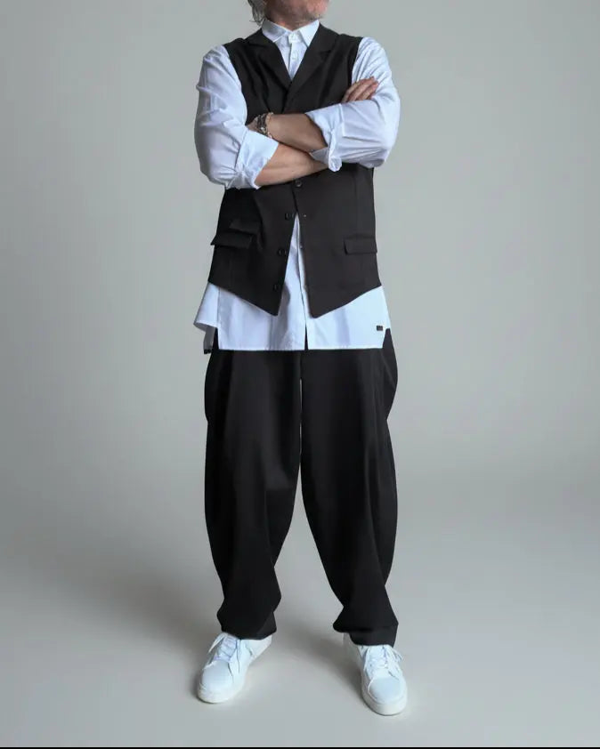 Model in baggy black pants, white shirt and black waistcoat standing in front of light grey backdrop