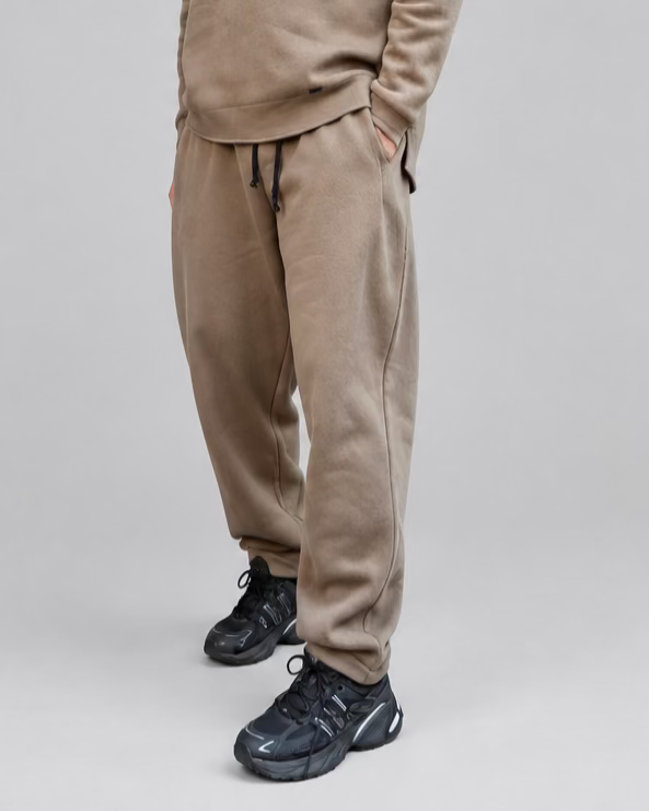 A man wearing a matching dark beige sweatshirt and jogger pants set, black sneakers, and sunglasses, standing in front of a black metal door with white brick walls.