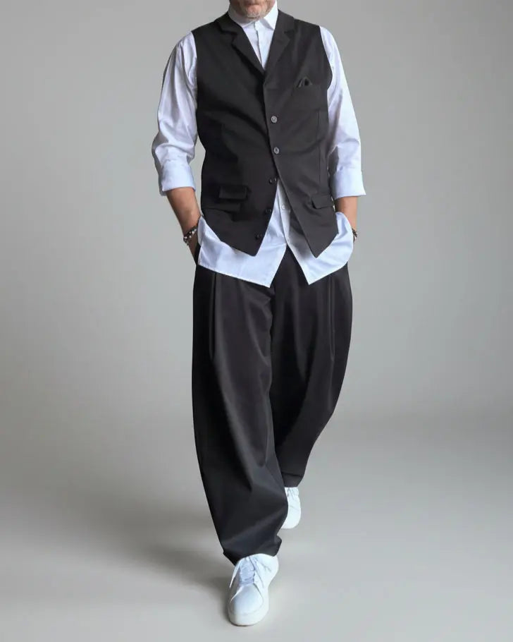 Model in baggy black pants, white shirt and black waistcoat standing in front of light grey backdrop