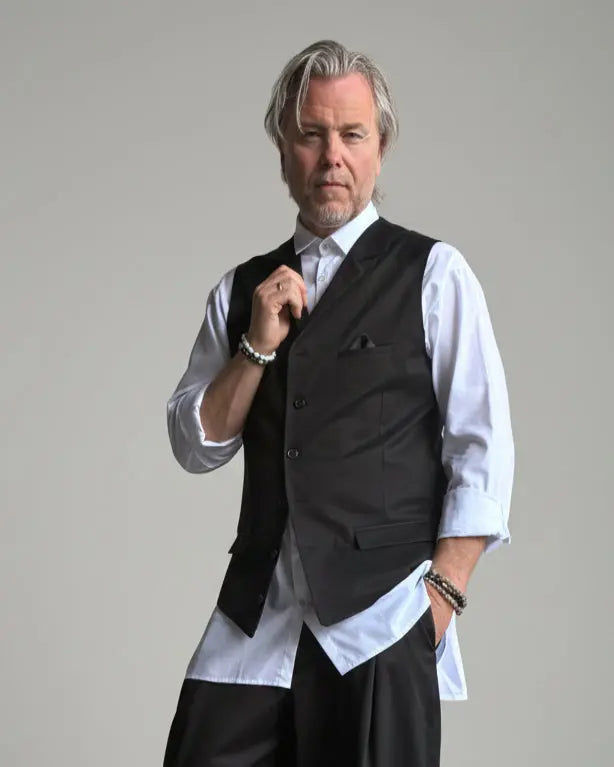 Model in baggy black pants, white shirt and black waistcoat standing in front of light grey backdrop
