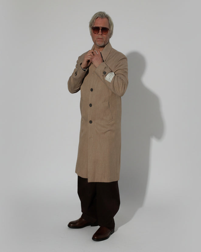 MODEL WEARING A BEIGE COAT IN FRONT OF A GREY BACKDROP