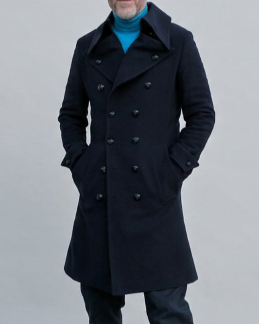 A man wearing a blue double-breasted coat with black metal buttons over a bright blue turtleneck, dark trousers, and brown leather shoes, standing against a light blue wall.