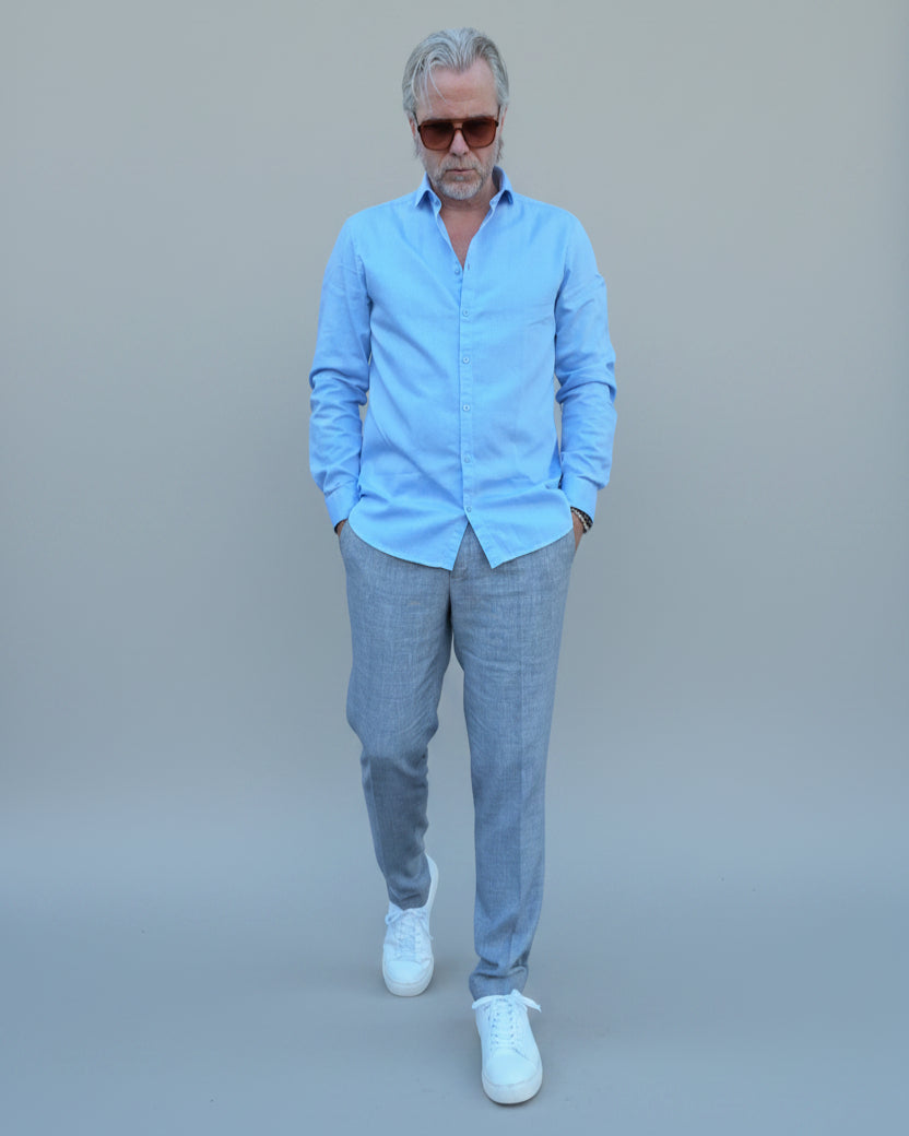 Man in a gray, blue checked pants and light blue shirt standing in front of a light grey backdrop.