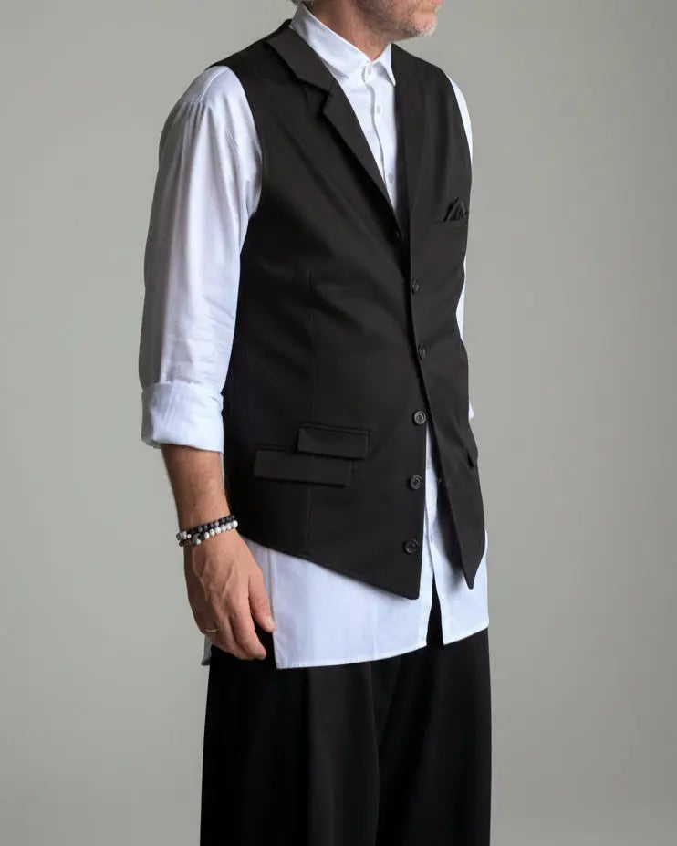 Model in baggy black pants, white shirt and black waistcoat standing in front of light grey backdrop