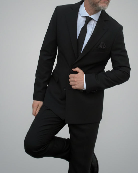 Model leaning against light grey backdrop wearing black suit and white shirt.